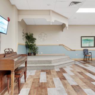 A spacious room with a piano, stage, and seating area, featuring wooden flooring and decorative plants.