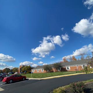 A clear blue sky adorned with fluffy clouds, overlooking a well-maintained landscape and parked cars.
