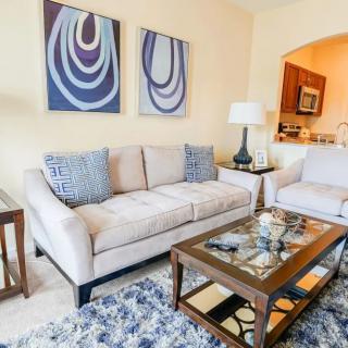 A cozy living room with a beige sofa, armchair, and a glass coffee table, accented by modern artwork and a patterned rug.