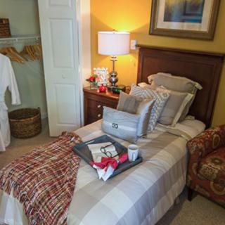 A cozy bedroom featuring a neatly made single bed with a tray of breakfast, a table lamp on the nightstand, and a robe hanging in the open closet.