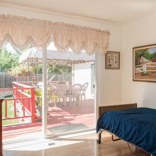 A cozy bedroom with a single bed, featuring a sliding glass door that opens to a sunlit deck with outdoor furniture.