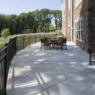 A sunny outdoor patio with a table and chairs, surrounded by a railing and trees.