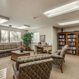 A cozy waiting room with patterned sofas, a wooden table, a bookshelf filled with books, and a potted plant.