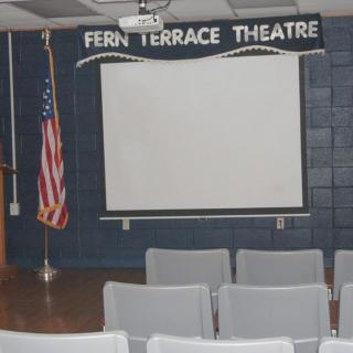 An indoor theater setup with chairs facing a screen, featuring a stage and an American flag.
