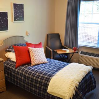 A cozy bedroom with a single bed adorned with red and white pillows, next to a window and a chair.