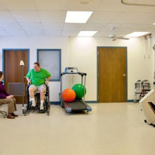 A patient in a wheelchair engages with a healthcare professional in a rehabilitation setting, surrounded by exercise equipment.