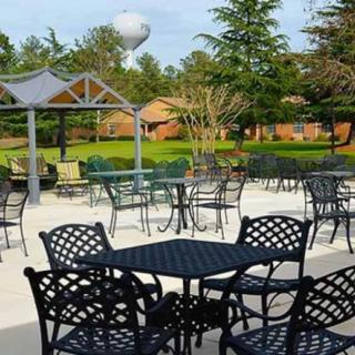 An outdoor seating area with metal tables and chairs, set in a park-like environment with greenery and trees.