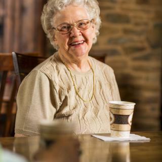 An elderly woman smiles while holding a coffee cup indoors.