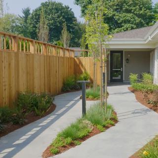 A landscaped garden pathway leads to a house entrance, bordered by a wooden fence and lush greenery.