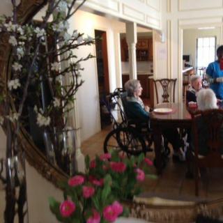 A cozy dining room with a group of people, including an elderly person in a wheelchair, gathered around a table. A mirror and flowers add warmth to the ambiance.
