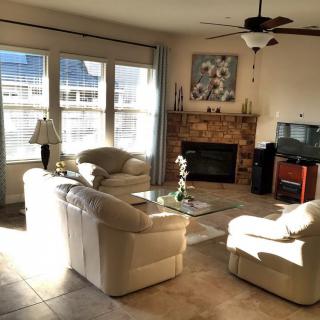 A cozy living room with sunlight streaming through large windows, featuring comfortable sofas, a fireplace, and a television.