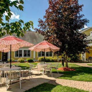 A charming outdoor patio area with tables and chairs shaded by red umbrellas, surrounded by trees and a yellow building.
