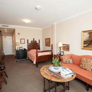 A cozy studio apartment with a combined living and sleeping area, featuring a sofa, round table, bed, and decorative elements like artworks and plants.