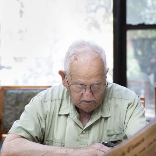 An elderly man with glasses is engrossed in reading a book indoors, bathed in natural light.