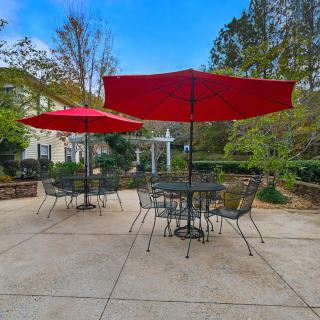 Outdoor patio area with tables, chairs, and red umbrellas surrounded by greenery.