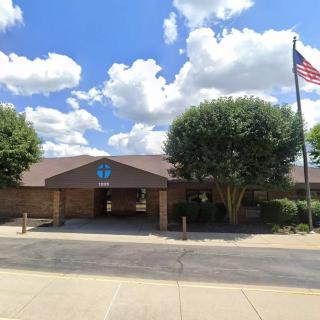 A one-story brick building with an American flag in front, surrounded by trees under a partly cloudy sky.