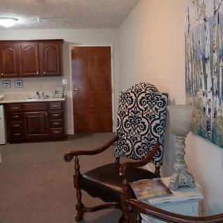 A cozy living space featuring a patterned chair, artwork on the wall, and a kitchenette in the background.
