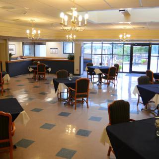 A spacious restaurant interior with neatly arranged tables and chairs, illuminated by chandeliers and natural light from large windows.