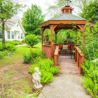 A charming gazebo surrounded by lush greenery and a small statue, with a house visible in the background.