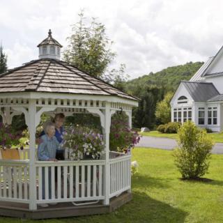 A scenic view of a white gazebo surrounded by lush greenery and flowers, with a house in the background.