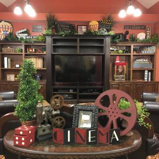 A cozy home theater setup with cinema-themed decor, featuring a vintage film reel, bookshelves, and greenery around a television.