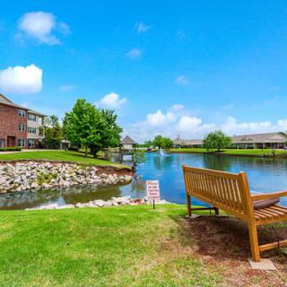 A serene lakeside view with a wooden bench, surrounded by houses and lush greenery under a bright blue sky.