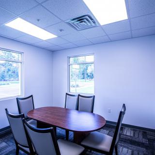 A small conference room with an oval table and six chairs, illuminated by overhead lighting and natural light from two windows.
