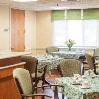A cozy dining area with neatly set tables and chairs, featuring floral tablecloths and a vase of flowers by the window.