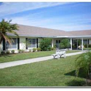 A single-story building with a well-maintained lawn, palm tree, and bench under a clear sky.