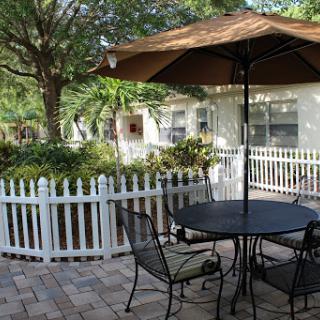 A cozy outdoor patio setup featuring a table and chairs under a shaded umbrella, surrounded by lush greenery and a white picket fence.