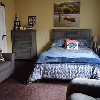 A cozy bedroom with a bed, dresser, and armchair, featuring a landscape painting on the wall.