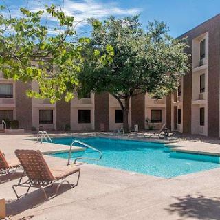 A serene outdoor swimming pool area surrounded by an apartment complex, featuring lounge chairs and lush greenery under a clear blue sky.