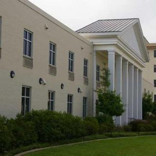 A building with classical columns and multiple windows, surrounded by a green lawn and trees.
