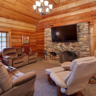 A cozy log cabin living room featuring a stone fireplace, comfortable seating, and a large television.