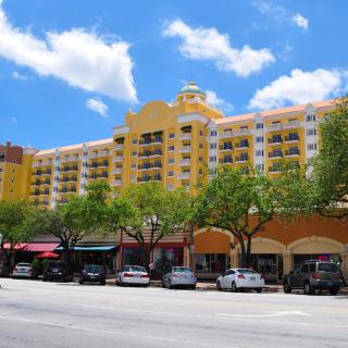 A view of a colorful residential building with shops below, lined with parked cars and trees on a sunny day.
