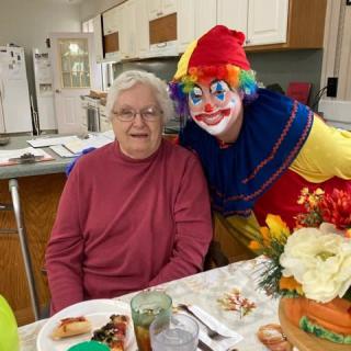 An elderly woman sitting at a dining table beside a smiling clown in a colorful costume.