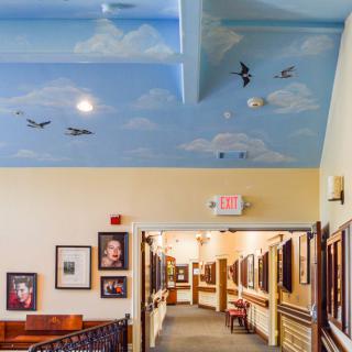 A hallway with a blue sky ceiling mural featuring clouds and birds, adorned with framed paintings along the walls.