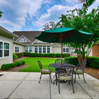 A cozy outdoor patio area with a table, chairs, and an umbrella surrounded by greenery.