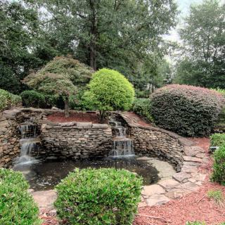 A serene garden scene featuring a small waterfall flowing into a pond, surrounded by lush greenery and stone paths.