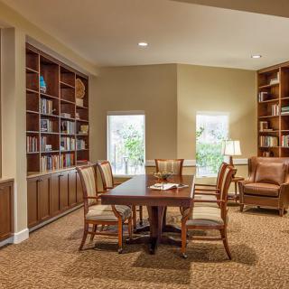 A cozy library space with bookshelves, a central table surrounded by chairs, and warm lighting.