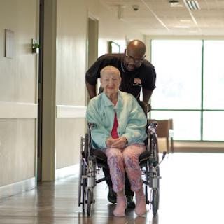 A caregiver gently pushes an elderly woman in a wheelchair down a bright hospital hallway.
