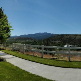 A scenic view featuring mountains in the background, a white fence along a pathway, and lush green grass under a clear blue sky.