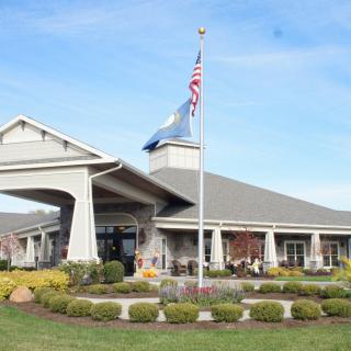 The image shows a large building with a sloped roof, featuring two flags flying in front and surrounded by neatly landscaped gardens.