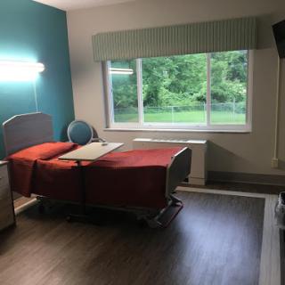 A well-lit hospital room featuring a patient bed, a chair, and a window with a view of greenery.
