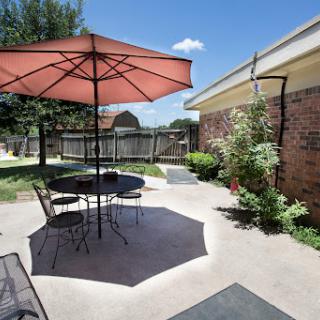 A sunny patio area featuring a large umbrella over a table with chairs, surrounded by greenery.