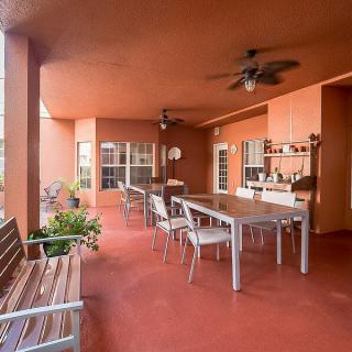 A spacious patio with outdoor seating and dining area under a terracotta-colored roof, featuring plants and fans for comfort.