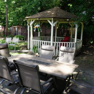 A tranquil garden scene featuring a gazebo surrounded by lush greenery and outdoor dining furniture on a patio.
