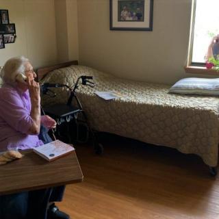 An elderly woman sits thoughtfully in a nursing home room with a bed and window.