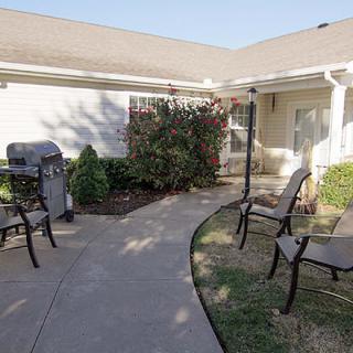 A cozy outdoor patio area with chairs, a grill, and a garden pathway in front of a house.