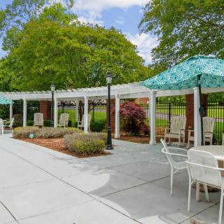 A serene patio area with a pergola, surrounded by lush greenery and furnished with tables and chairs.